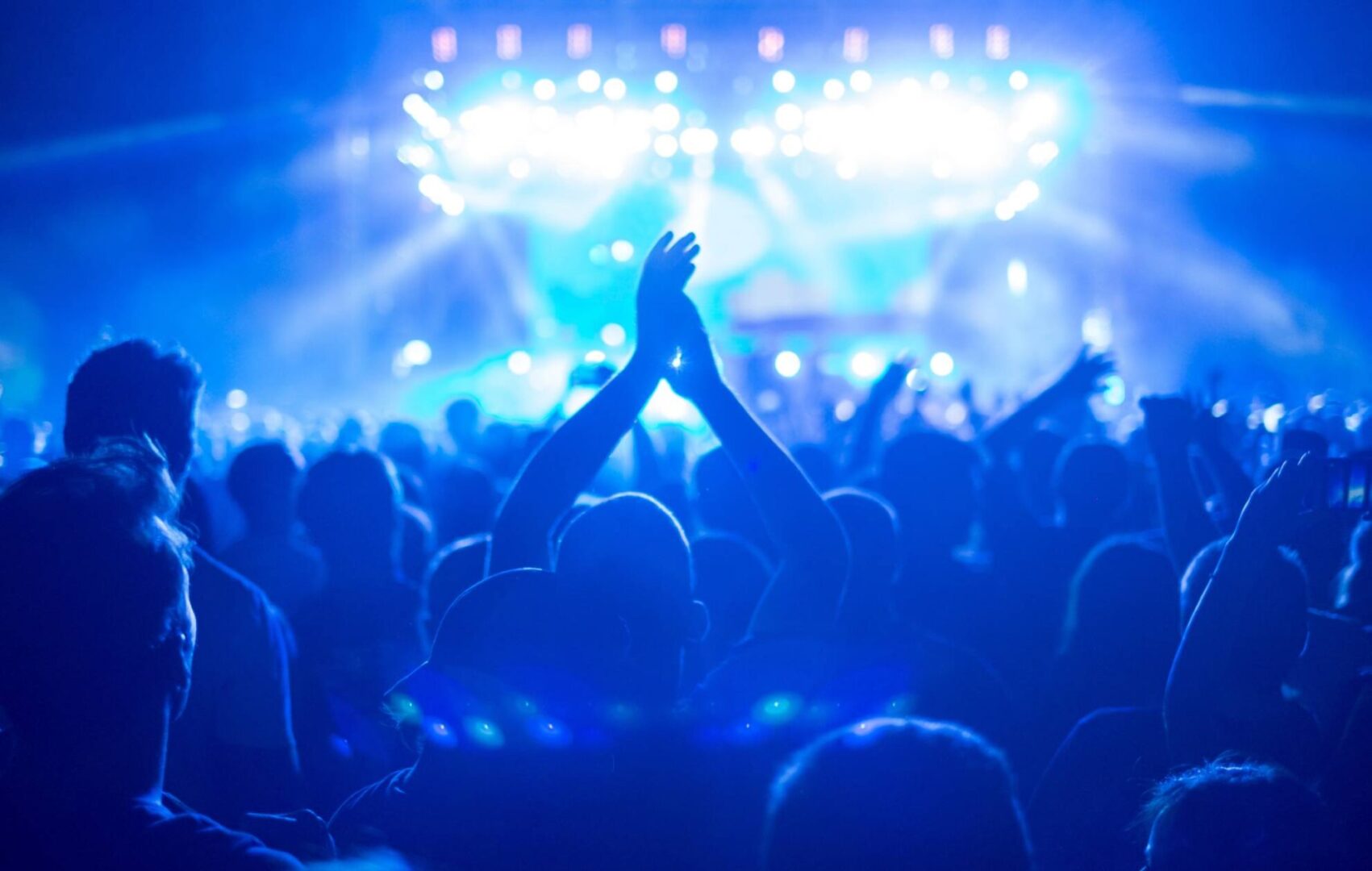 Shot of a large crowd at a music concert, enjoying a music festival event, with blue stage lights, sea of hands. Credit: zoramn via GETTY